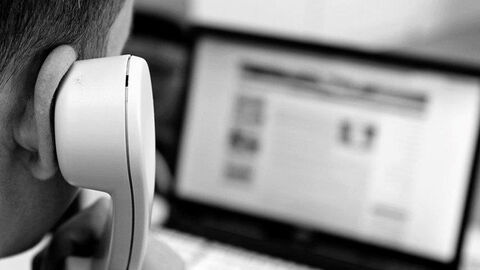 Eine von hinten aufgenommene Person hält einen Telefonhörer am Ohr, im Hintergrund ist ein Bildschirm eines Computers