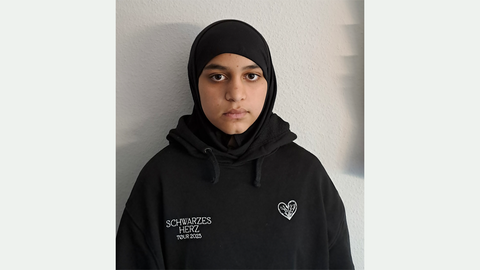 Eine 13-Jährige mit dunklen Augen, schwarzem Kopftuch und schwarzem Kapuzenpullover mit der weißen Aufschrift SCHWARZES HERZ TOUR 2023 und weißes eingesticktes Herz