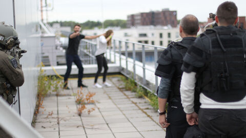 Im Hintergrund wird eine Person mit einem Messer bedroht, im Vordergrund sind Beamte der Verhandlungsgruppe von hinten zu sehen