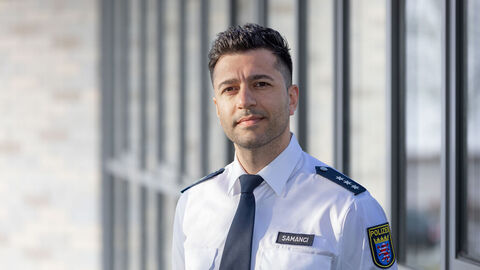 Migrationsbeauftragter Berkant Samanci in Polizeiuniform vor Glaswand stehend 