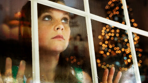 Mädchen schaut traurig aus dem Fenster, im Hintergrund Weihnachtsbeleuchtung 